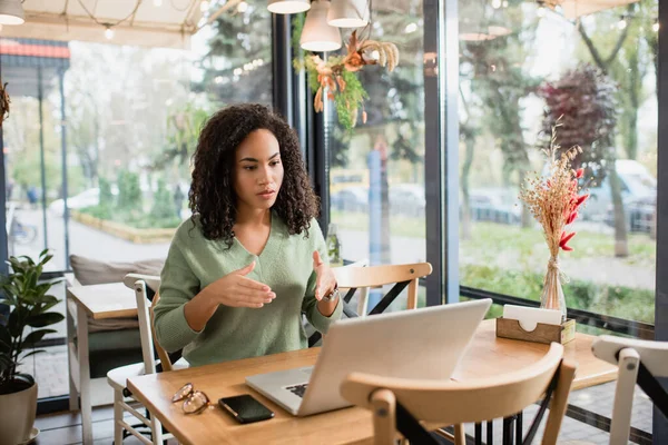 Curly african american woman gesturing while explaining and looking at laptop during video call — Stock Photo