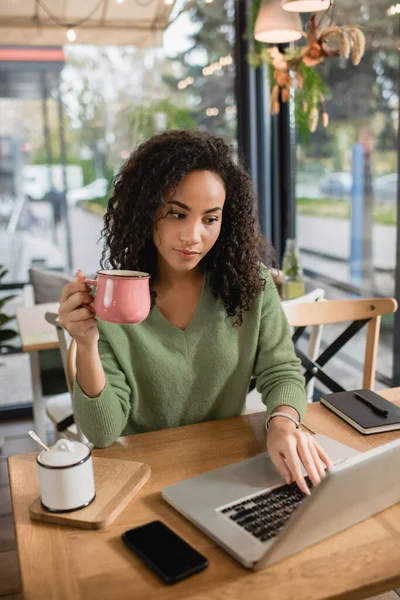 Mujer afroamericana sosteniendo taza de café mientras usa el ordenador portátil en la cafetería - foto de stock