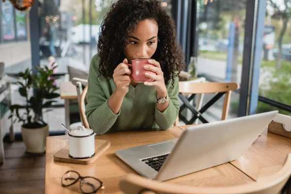 Afrikanerin trinkt Kaffee aus Tasse, während sie auf Laptop schaut — Stockfoto