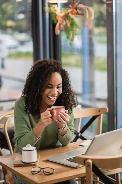 Joyful african american woman holding cup while looking at laptop in cafe — Stock Photo