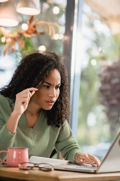 Mujer afroamericana enfocada sosteniendo pluma y mirando a la computadora portátil mientras estudia en línea en la cafetería - foto de stock