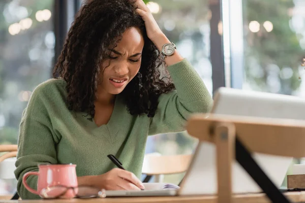 Épuisé femme afro-américaine écrit dans un cahier tout en étudiant en ligne dans un café — Photo de stock