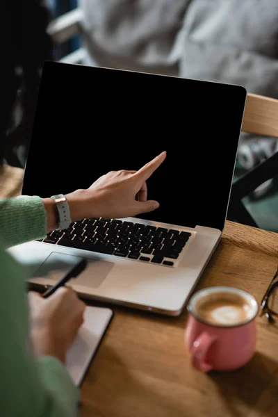 Vista parcial de la mujer afroamericana señalando con el dedo a la computadora portátil con pantalla en blanco - foto de stock