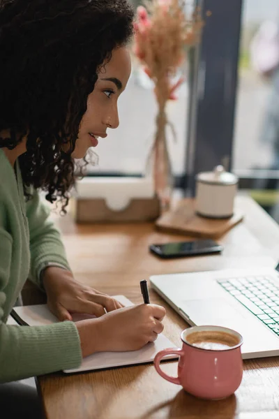 Donna afro-americana che scrive nel taccuino vicino alla tazza di latte mentre studia online nel caffè — Foto stock