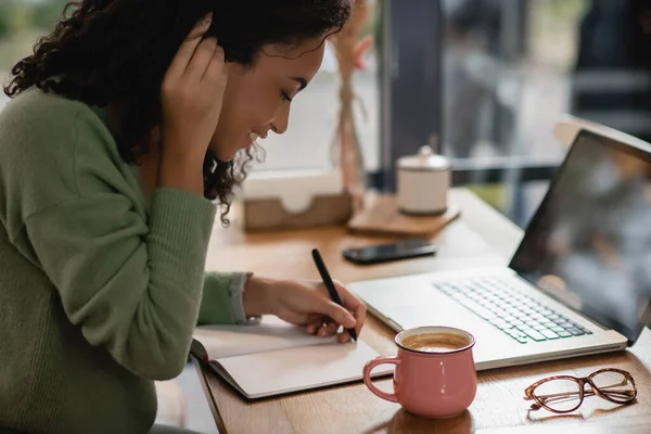 Felice donna afroamericana che fissa i capelli mentre scrive nel notebook vicino al laptop e alla tazza di caffè nel caffè — Foto stock