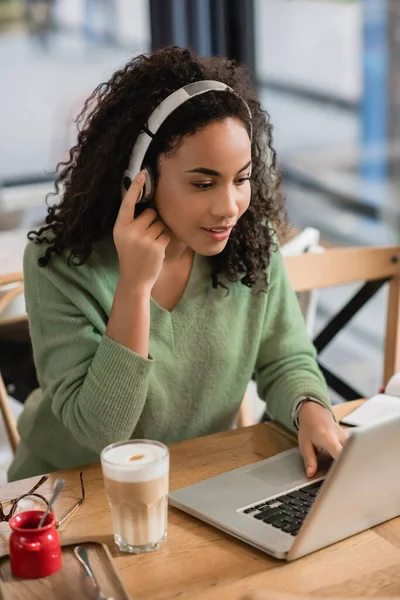 Mujer afroamericana escuchando podcast cerca del ordenador portátil en la cafetería - foto de stock