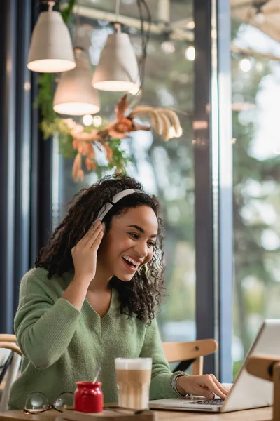 Mujer afroamericana feliz escuchando podcast cerca de la computadora portátil en la cafetería - foto de stock