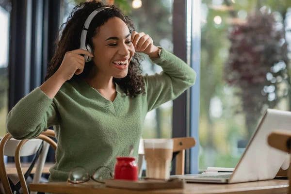 Joyeuse femme afro-américaine écoutant podcast près d'un ordinateur portable dans un café — Photo de stock