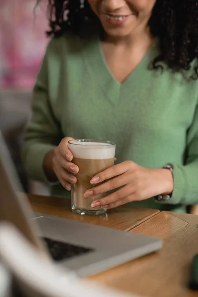 Cropped view of african american woman holding glass of latte near laptop on blurred foreground — Stock Photo