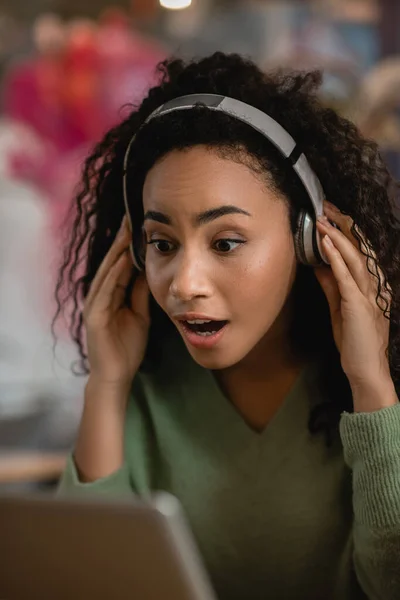 Mujer afroamericana sorprendida escuchando podcast cerca de la computadora portátil en la cafetería - foto de stock