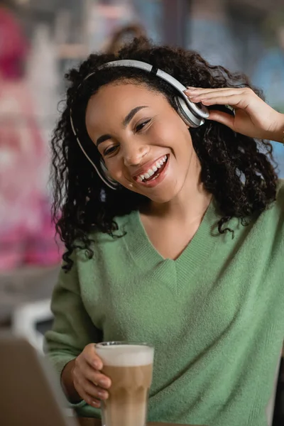 Mujer afroamericana feliz escuchando podcast y sosteniendo un vaso de café con leche en la cafetería - foto de stock