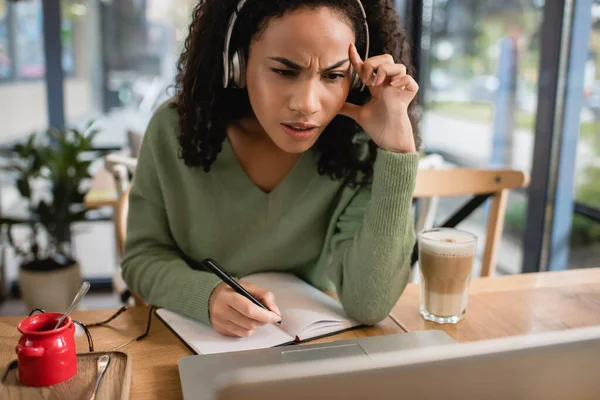 Зосереджена афроамериканська жінка слухає подкаст і дивиться на ноутбук в кафе — стокове фото