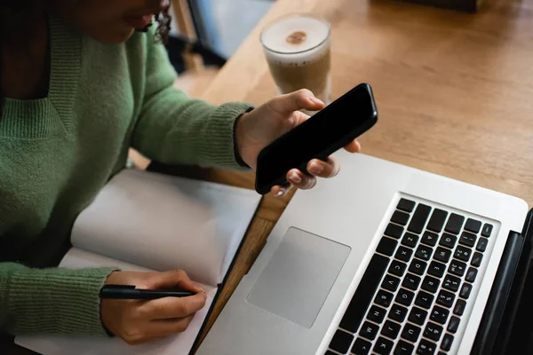 Обрізаний вид афроамериканки, що тримає смартфон з порожнім екраном біля ноутбука і ноутбука — стокове фото