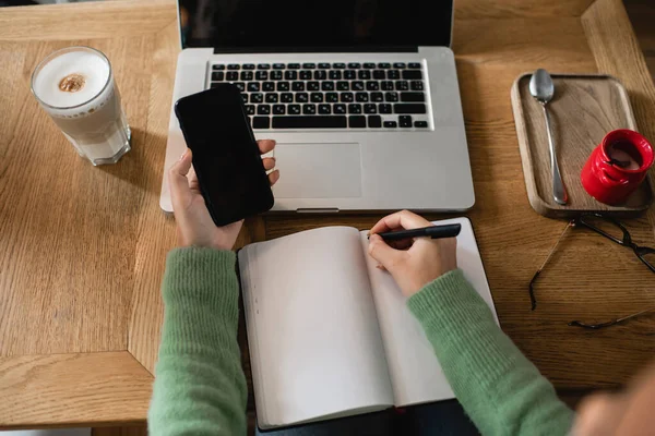 Vista recortada de la mujer afroamericana sosteniendo teléfono inteligente y pluma cerca de la computadora portátil, vaso de café con leche y portátil - foto de stock