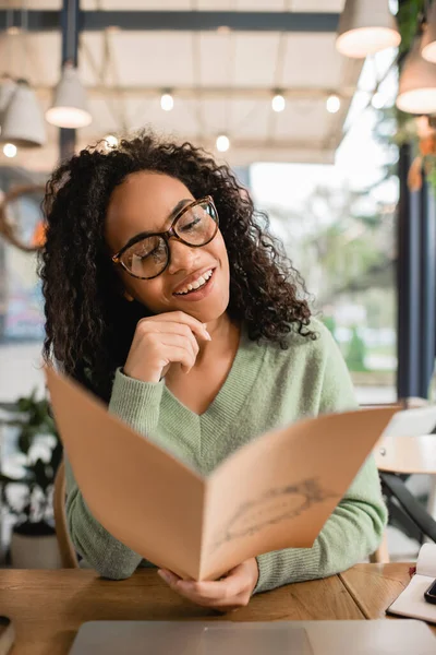 Feliz rizado africano americano mujer en gafas celebración menú en borrosa primer plano - foto de stock