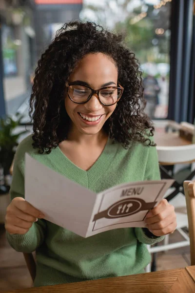 Joyeuse femme afro-américaine bouclée dans des lunettes tenant le menu au premier plan flou — Photo de stock