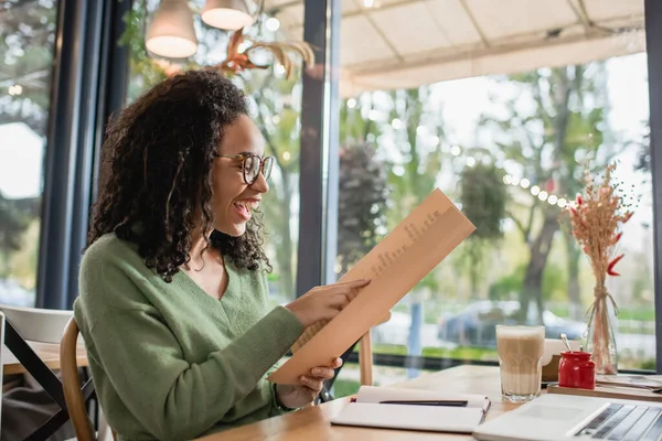 Femme afro-américaine gaie dans les lunettes en regardant le menu près de l'ordinateur portable dans le café — Photo de stock