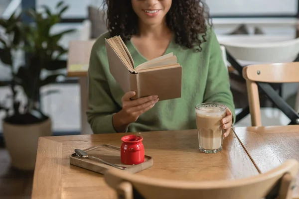 Abgeschnittene Ansicht einer glücklichen afrikanisch-amerikanischen Frau, die im Café Buch liest — Stockfoto