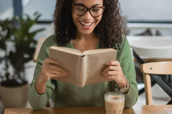 Alegre afroamericana mujer leyendo libro cerca de vidrio con café con leche en la cafetería - foto de stock