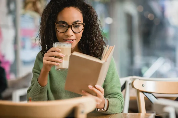 Mulher afro-americana alegre ler livro e segurando vidro com latte no café — Fotografia de Stock