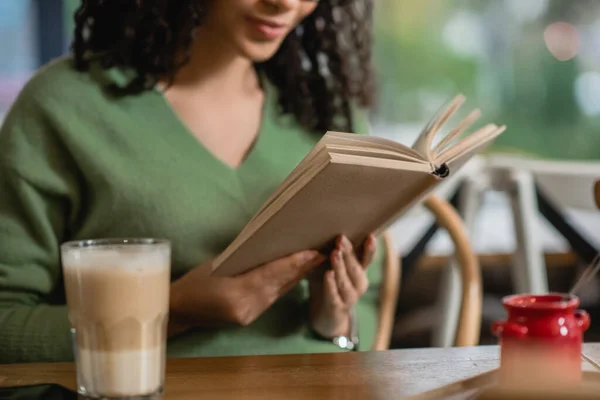 Vue partielle d'une femme afro-américaine lisant un livre près du verre avec du latte au premier plan flou — Photo de stock