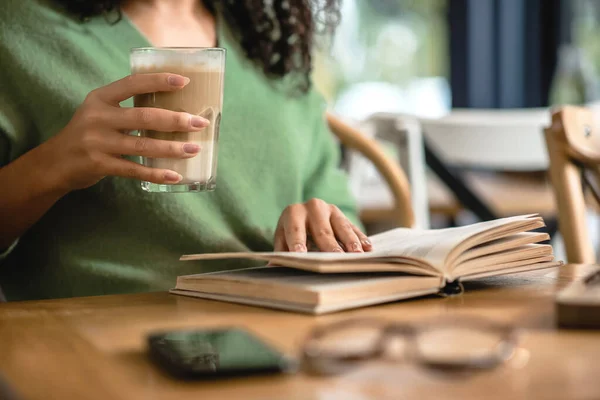 Vista ritagliata di donna afro-americana in possesso di vetro con latte durante la lettura di libro in caffè — Foto stock