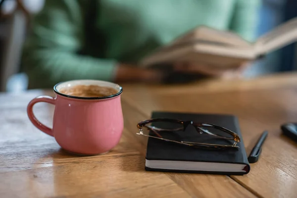 Mug with coffee near eyeglasses, notebook and african american woman on blurred background — Stock Photo