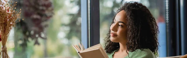 Mujer afroamericana mirando lejos cerca de libro en la cafetería, pancarta - foto de stock