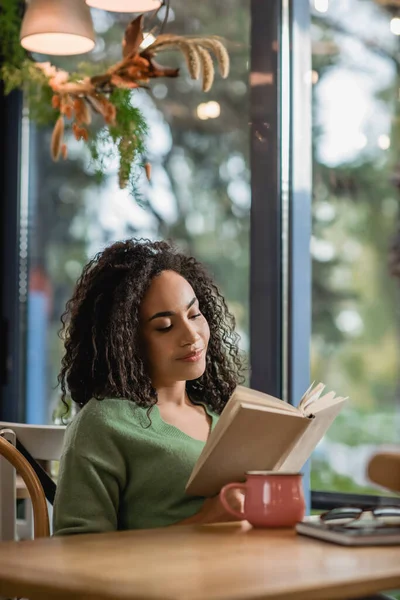 Curly african american woman reading novel in cafe — Stock Photo