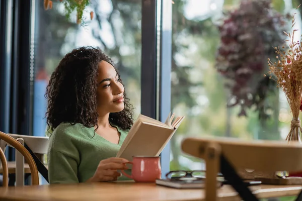 Verträumte afrikanisch-amerikanische Frau hält Buch in der Hand und blickt auf Fenster im Café — Stockfoto