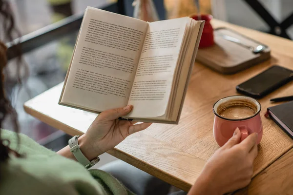 Visão parcial da mulher afro-americana segurando livro e xícara de cappuccino no café — Fotografia de Stock