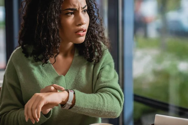 Preocupante mulher afro-americana tocando relógio no pulso enquanto espera no café — Fotografia de Stock
