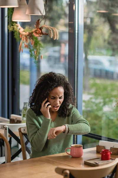 Расстроен африканский американец женщина разговаривает на смартфоне во время проверки часы на запястье и ожидания в кафе — стоковое фото