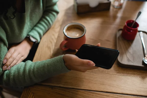 Vista parcial de la mujer afroamericana sosteniendo teléfono inteligente con pantalla en blanco cerca de la taza de capuchino - foto de stock