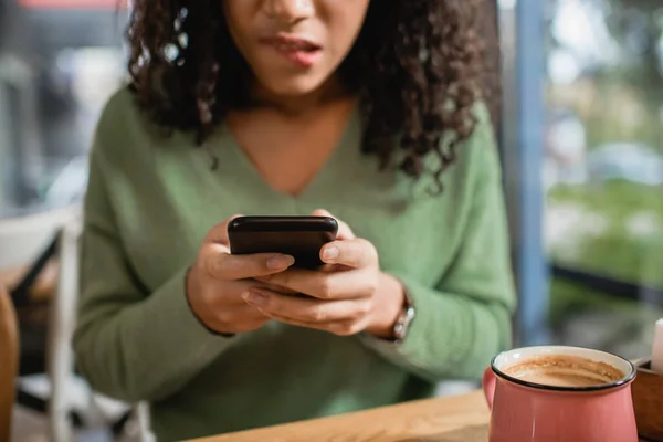 Taza de café con leche cerca preocupada mujer afroamericana mordiendo los labios mientras que los mensajes de texto en el teléfono inteligente en primer plano borrosa - foto de stock