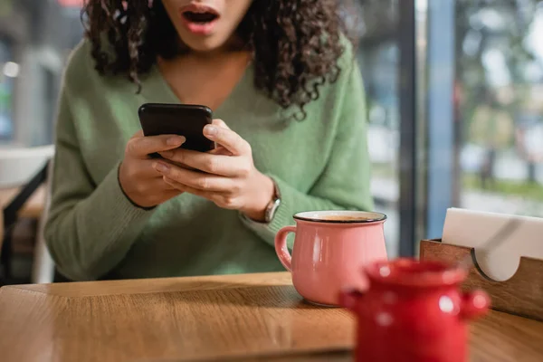 Xícara de café perto de mulher afro-americana chocada mensagens de texto no smartphone em primeiro plano desfocado — Fotografia de Stock