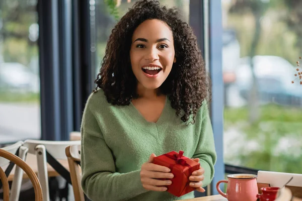 Amazed young african american woman holding red christmas gift box in cafe — Stock Photo