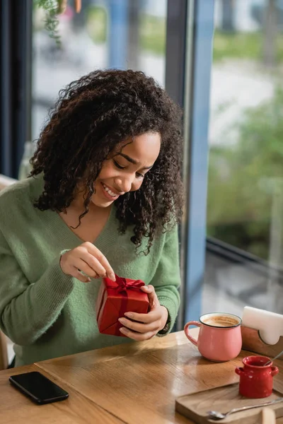 Mujer afroamericana feliz tirando de la cinta en la caja de regalo de Navidad roja en la cafetería - foto de stock