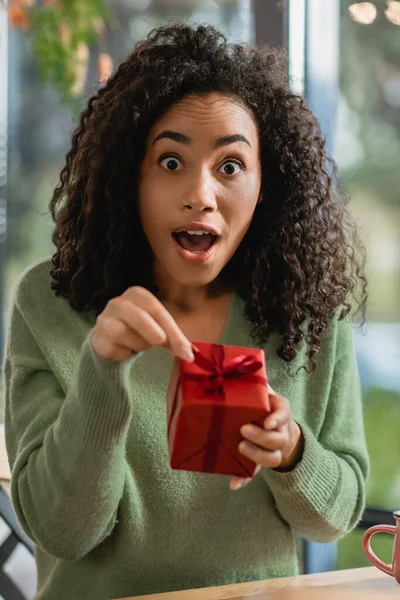 Shocked african american woman pulling ribbon on red christmas gift box in cafe — Stock Photo