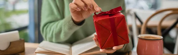 Vista ritagliata della donna afro-americana tirando nastro sulla scatola regalo di Natale rosso in caffè, banner — Foto stock