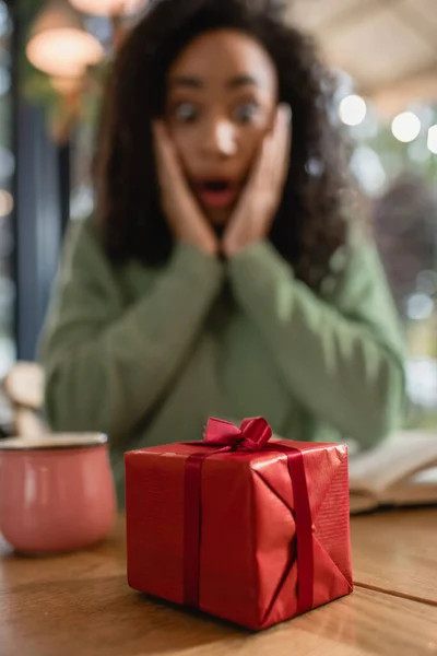 Rote Weihnachtsgeschenkschachtel in der Nähe schockierter Afroamerikanerin auf verschwommenem Hintergrund — Stockfoto