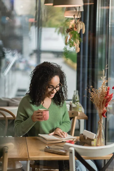 Mulher africana americana em óculos segurando xícara de café e livro de leitura no café — Fotografia de Stock