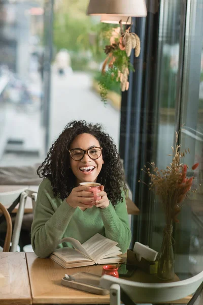 Donna afro-americana stupita negli occhiali che tengono la tazza di caffè vicino al libro sul tavolo nel caffè — Foto stock