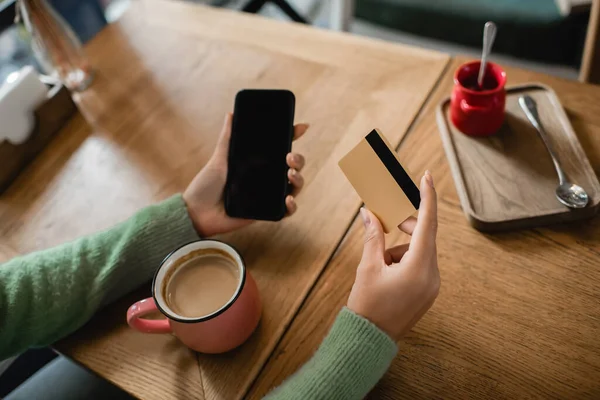 Vista ritagliata di donna afro-americana in possesso di carta di credito vicino smartphone con schermo bianco e tazza sul tavolo — Foto stock