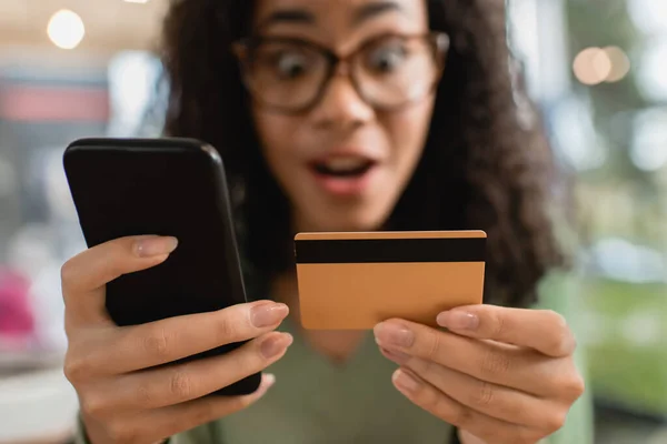 Smartphone et carte de crédit dans les mains de femme afro-américaine étonnée sur fond flou — Photo de stock