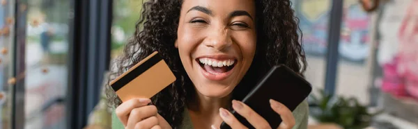 Cheerful african american woman laughing while holding smartphone and credit card, banner — Stock Photo