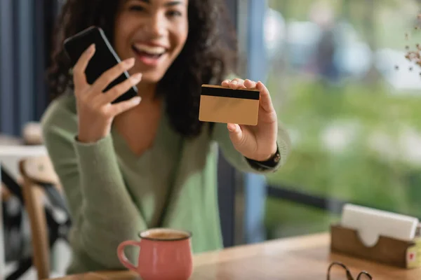 Tarjeta de crédito y teléfono inteligente en manos de la mujer afroamericana emocionada sobre fondo borroso - foto de stock