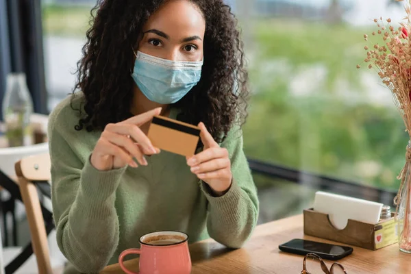 Африканская американка в медицинской маске держит кредитку возле смартфона и чашку на столе в кафе — стоковое фото