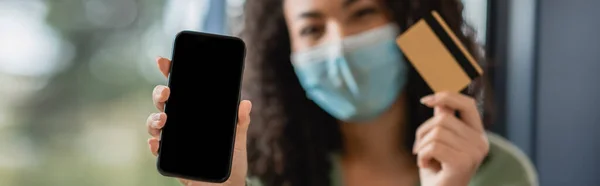 Smartphone com tela em branco e cartão de crédito em mãos de mulher afro-americana em máscara médica em fundo borrado, banner — Fotografia de Stock