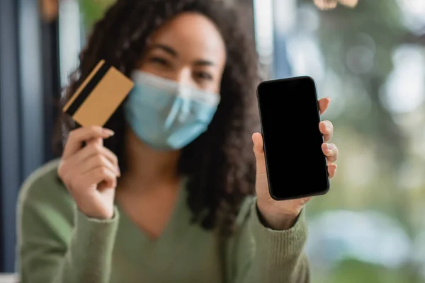 Smartphone con schermo bianco e carta di credito in mano di donna afroamericana in maschera medica su sfondo sfocato — Foto stock